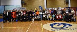 Zdjęcie grupowe uczestnicy, goście organizatorzy sędziów uczniowie z przodu klęczą wszyscy na płycie hali śremskiego sportu