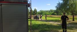 na pierwszym planie strażak w koszulce i czarnych spodniach z lewej strony widać kawałek samochodu strażackiego w oddali uczestnicy obozu wokół drzewa