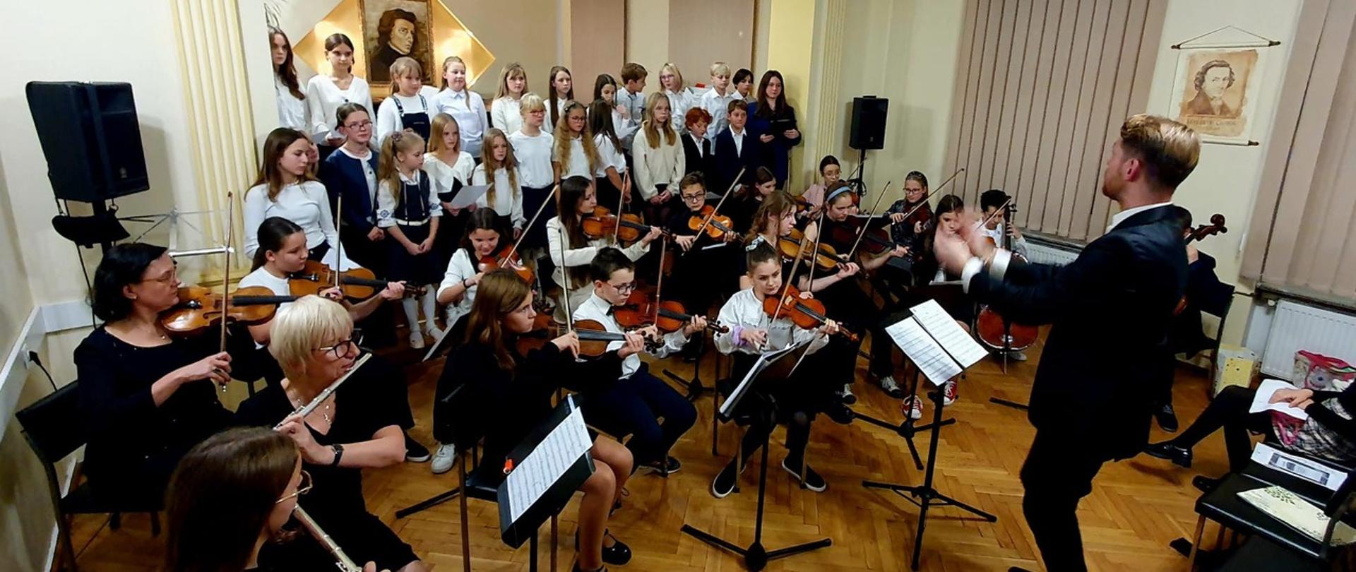 Pan Dominik Jarocki dyryguje chór i orkiestrę PSM I st. w Kłodzku, uczniowie w pierwszym rzędzie grają na instrumentach, na dalszym planie stoją śpiewając