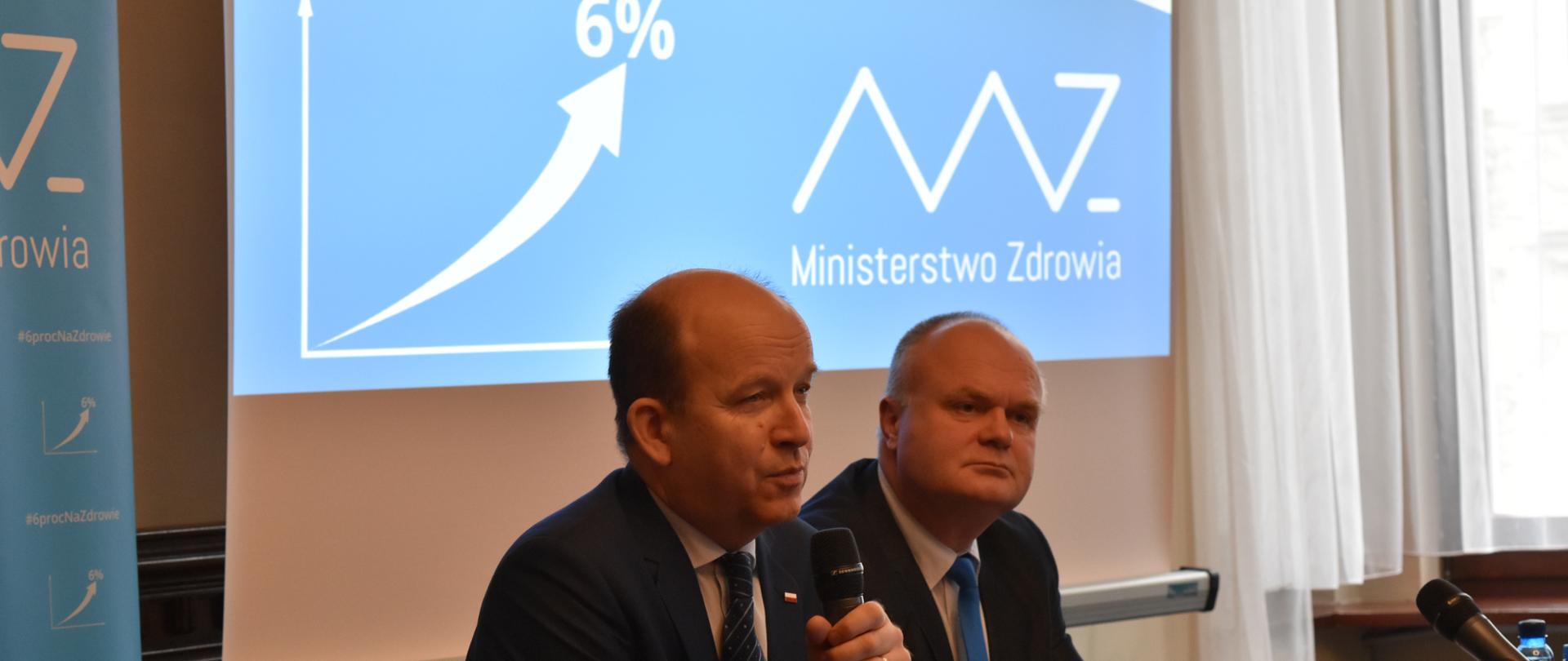 Minister Zdrowia Konstanty Radziwiłł siedzi przy stole i na tle prezentacji multimedialnej opowiada o wzroście nakładów na zdrowie do poziomu 6% PKB. 