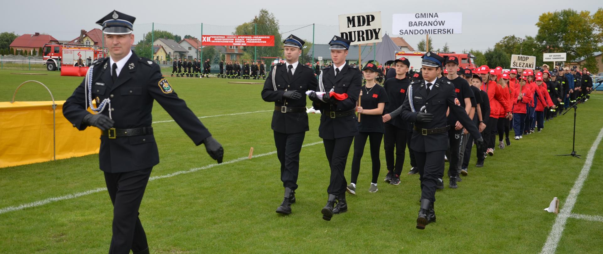 Strażacy w ugrupowaniu marszowym maszerują na płycie stadionu sportowego. Na czele pododdziału maszeruje trzech strażaków, z których jeden przenosi na ramionach flagę Rzeczypospolitej. Ugrupowanie prowadzi dowódca. 