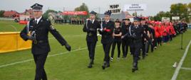 Strażacy w ugrupowaniu marszowym maszerują na płycie stadionu sportowego. Na czele pododdziału maszeruje trzech strażaków, z których jeden przenosi na ramionach flagę Rzeczypospolitej. Ugrupowanie prowadzi dowódca. 
