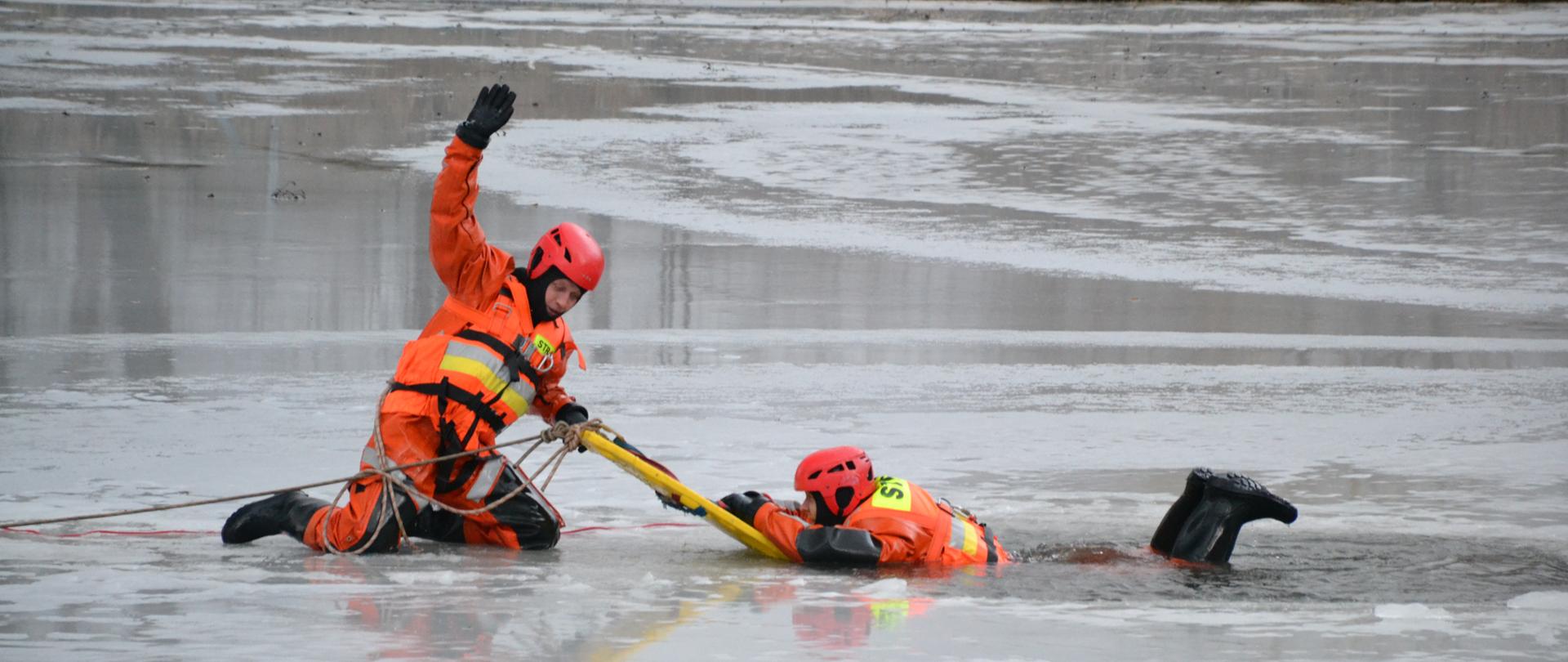 Dwóch ratowników w skafandrach, jeden zanurzony w przeręblu, drugi na tafli lodu podejmuje pierwszego z wody

