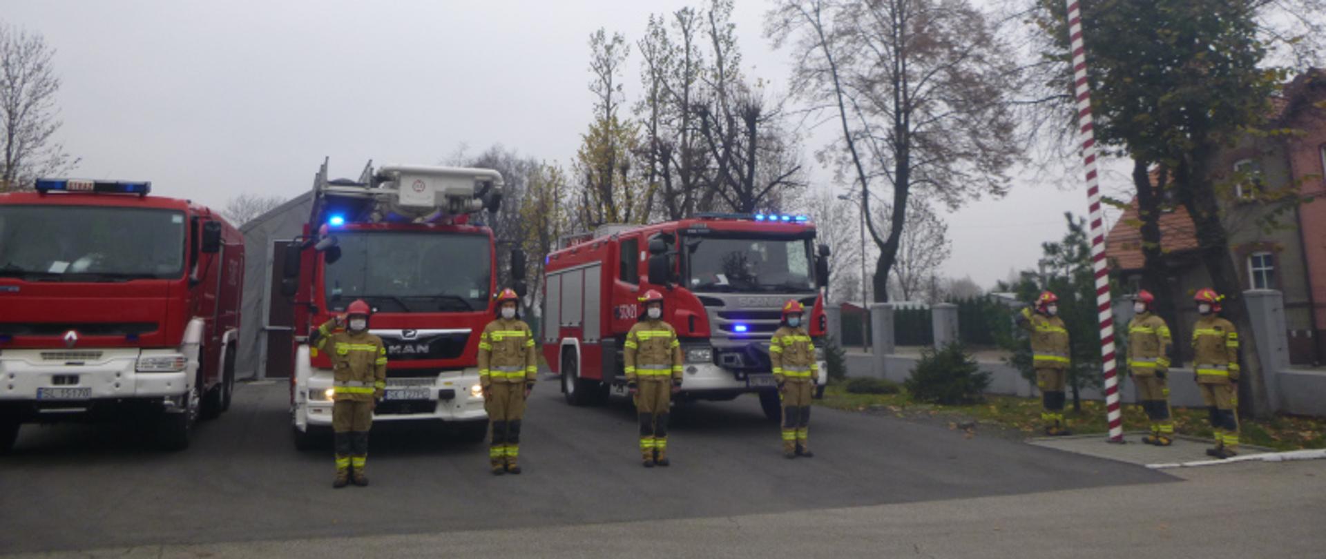 Na zdjęciu znajdują się strażacy w umundurowaniu specjalnym w czasie hymnu państwowego. W tle znajdują się samochody gaśnicze oraz podnośnik hydrauliczny. Trzech strażaków znajduje się przy macie flagowym, natomiast czterech strażaków ustawionych jest przed samochodami pożarniczymi.