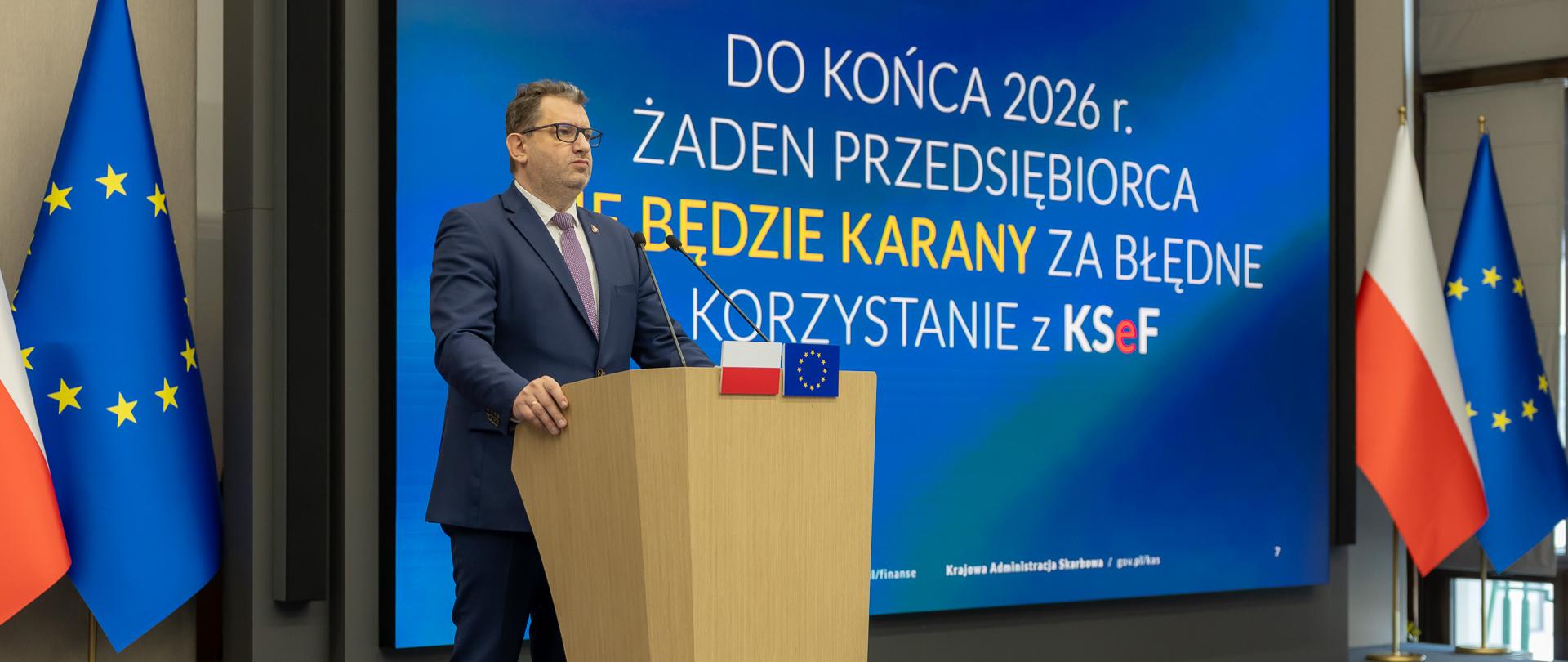 Marcin Łoboda, Sekretarz Stanu w Ministerstwie Finansów, Szef Krajowej Administracji Skarbowej, stoi za mównicą, w tle ekran z prezentacją o KSeF oraz flagi Polski i UE