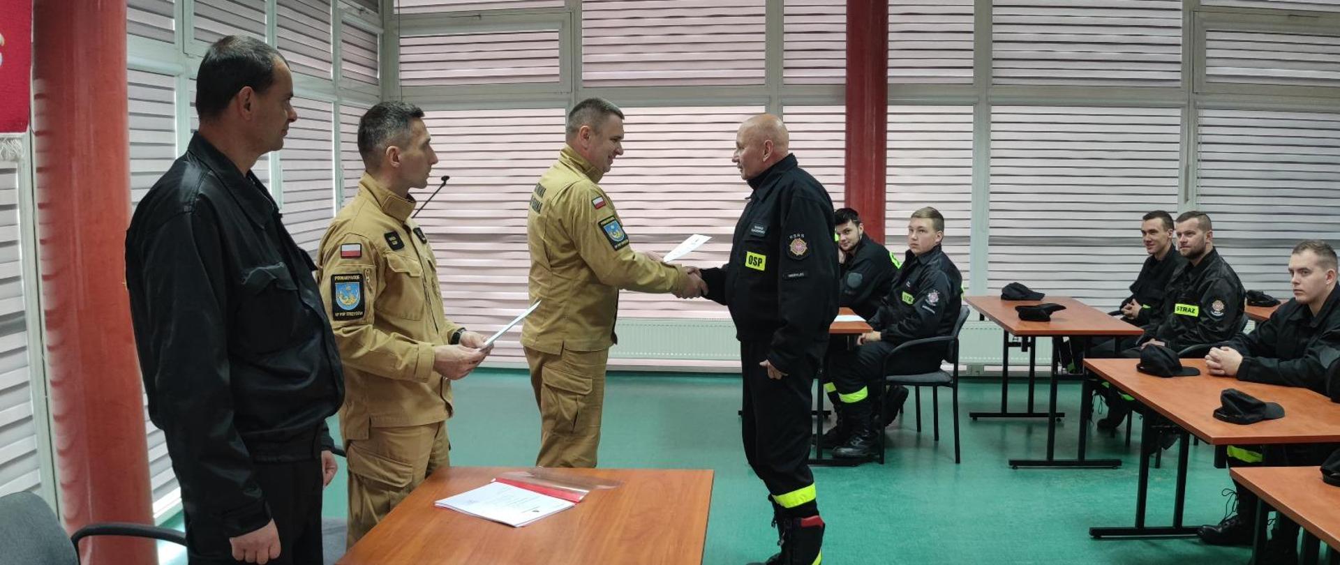 Grupa mężczyzn w mundurach, po lewej stronie stoją, dwóch ściska sobie dłonie, po prawej stronie siedzą w ławkach.