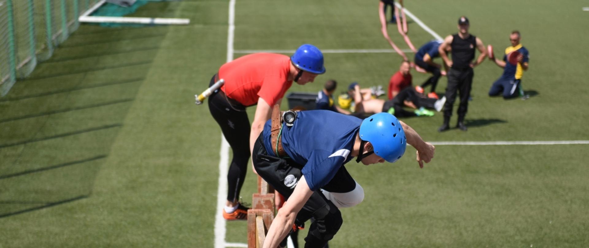 Dwóch mężczyzn przeskakuje przez przeszkodę nazywaną ściana. Ubrani są w strój sportowy. W tle boisko na którym ćwiczą zawodnicy. 