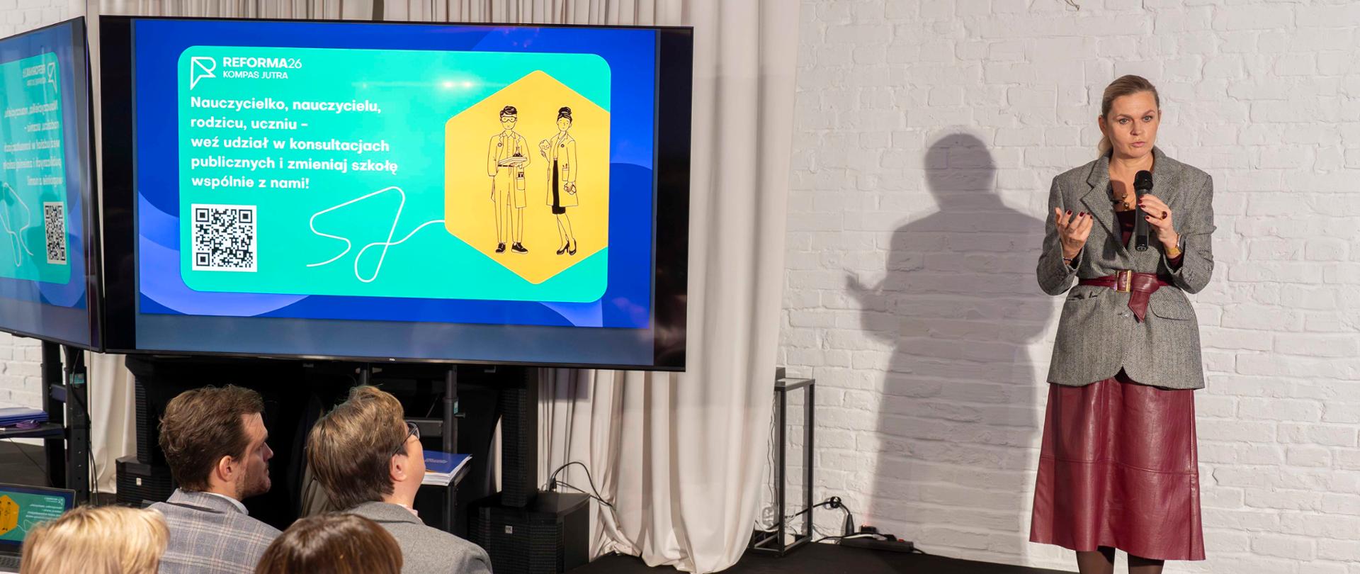 Kobieta w szarej, tweedowej marynarce i czerwonej spódnicy stoi na scenie i mówi do mikrofonu, gestykulując obiema rękami. Za nią widoczny jest duży ekran z napisem "REFORMA 26 KOMPAS JUTRA" i hasłem zachęcającym do udziału w konsultacjach publicznych.