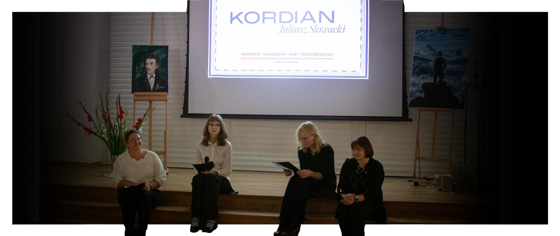 Nauczycielki Patrycja Cicha oraz Agnieszka Mierzwiak wraz z dwójką uczennic w trakcie Narodowego Czytania na auli w PLSP z ekranem w tle, na którym znajduje się napis KORDIAN Juliusz Słowacki. Po bokach obrazy przedstawiające Juliusza Słowackiego oraz Kordiana.