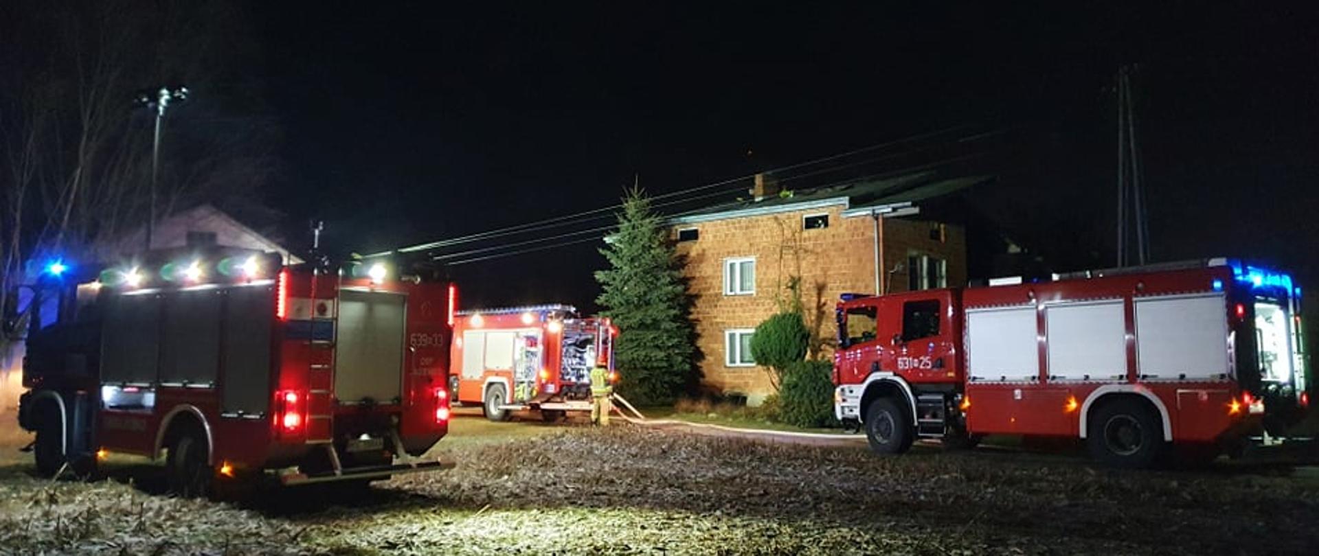 Zdjęcie wykonane o zmroku. Widoczne trzy samochody gaśnicze oraz dwukondygnacyjny budynek mieszkalny z czerwonej cegły, w którym były prowadzone działania.