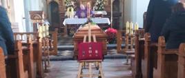 Pogrzeb strażaka. Kościół parafialny w Łaszczynie. Przy ołtarzu kapłan odprawia mszę św. w intencji Zmarłego. W nawie głównej stoi trumna, przed nią poduszka z przypiętymi odznaczeniami oraz wiązanki kwiatów.