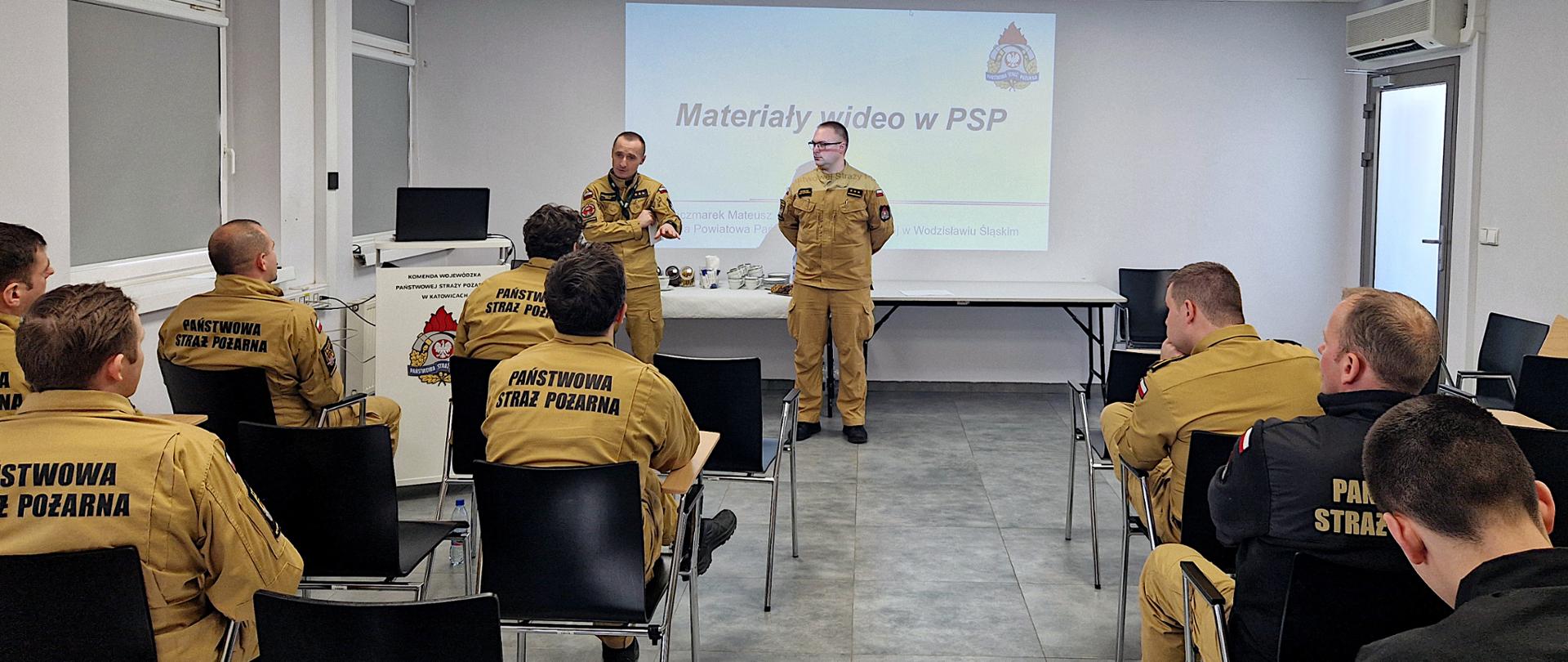 Na sali szkoleniowej siedzą odwróceni tyłem, w rzędach strażacy w piaskowych mundurach służbowych. Na wprost stoi dwóch strażaków, prowadzą wykład. Za nimi, na ścianie rzutnik wyświetla prezentację. Przy ścianie ustawione dwa stoliki.