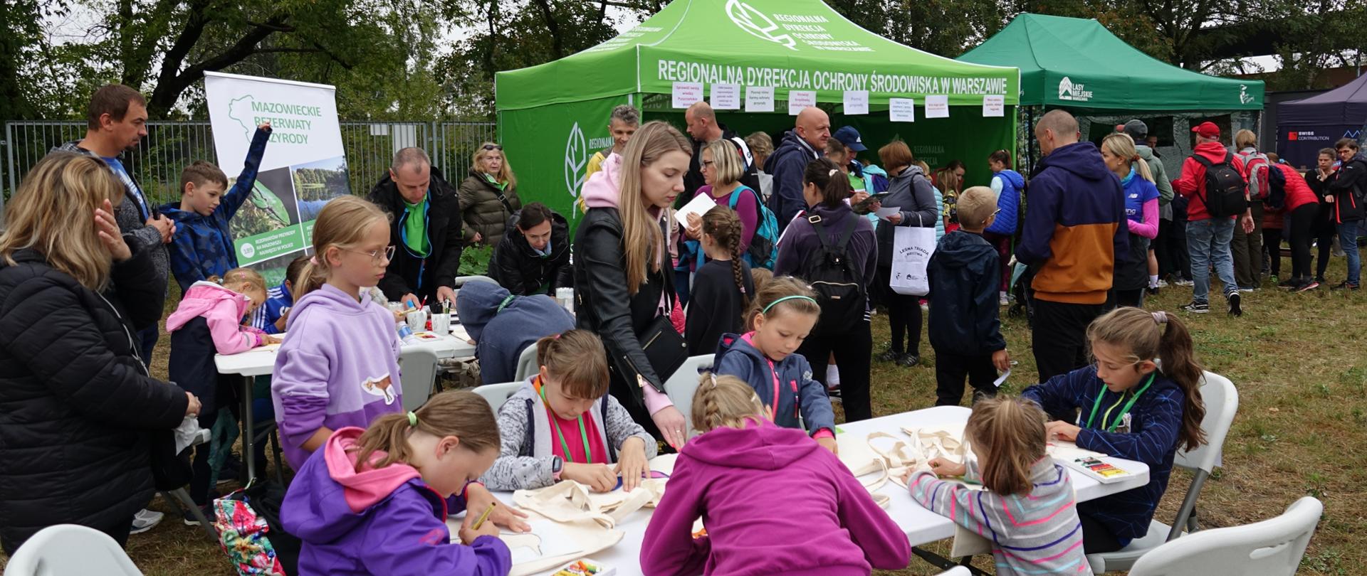 Festyn - na pierwszym planie dzieci przy stolikach wykonują prace plastyczne, w tle namiot i uczestnicy konkursów
