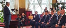 Minister przemawia do mikrofonu, stojąc przed salą pełną ludzi.
