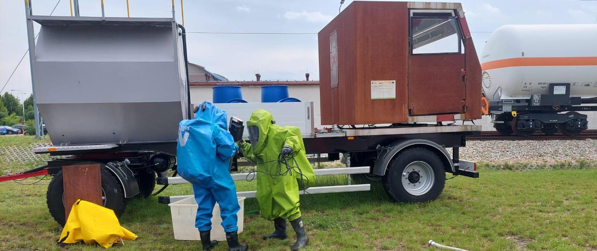 Poligon CS PSP, dwóch strażaków w specjalistycznych strojach ratownictwa chemicznego wykonuje działania przy makiecie samochodu przewożącego ładunek chemiczny