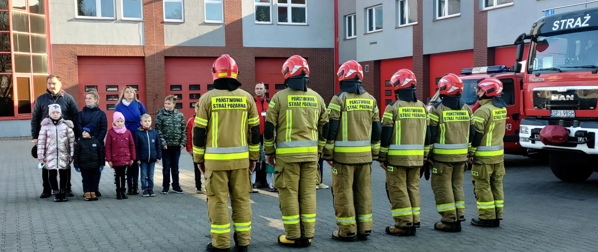 Zdjęcie przedstawia grupę strażaków stojących w szeregu. Na drugim planie widać grupę dzieci wraz z opiekunami. Z prawej strony stoją samochody ratowniczo - gaśnicze.