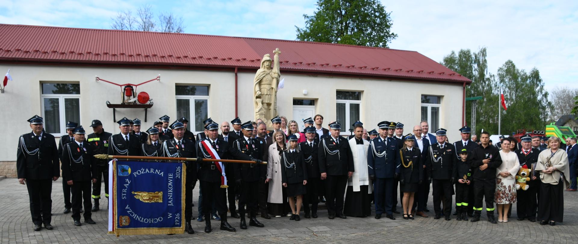 Grupowe zdjęcie uczestników uroczystości strażackiej przed budynkiem remizy. Wśród zgromadzonych znajdują się strażacy w galowych i bojowych mundurach, przedstawiciele lokalnej społeczności oraz duchowny, a na pierwszym planie widoczny jest sztandar Ochotniczej Straży Pożarnej