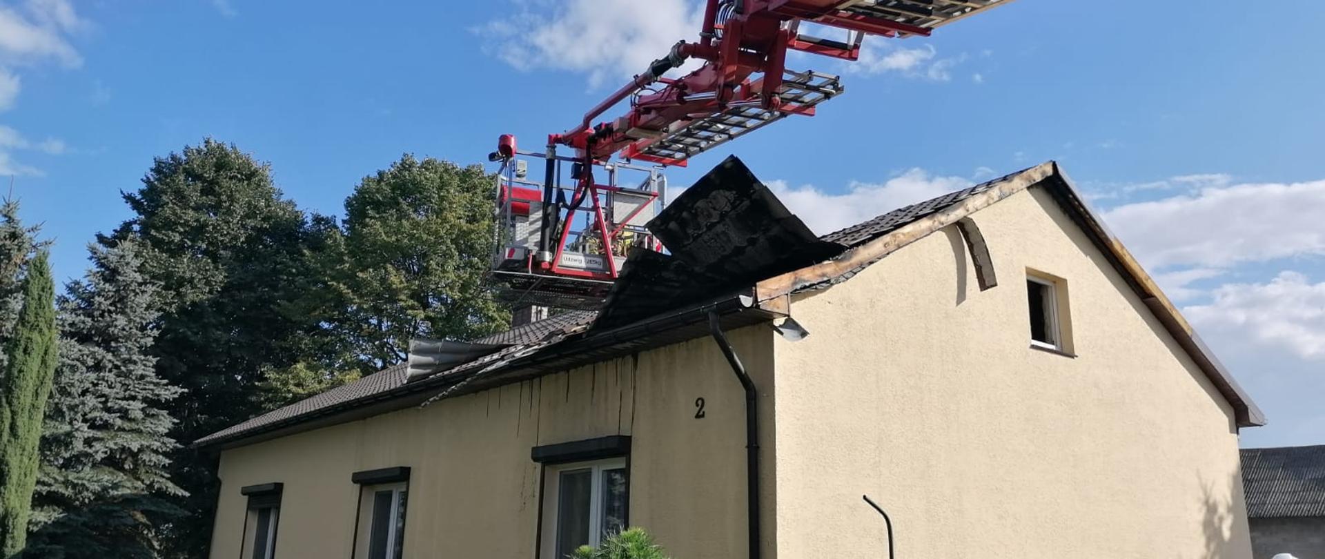 Budynek jednorodzinnym, parterowy, w kolorze żółtym, z dachem skośnym. Po prawej stronie ramię podnośnika PSP SHD 24. W koszy ratownik ubrany w ubranie specjalne koloru piaskowego. Niebo błękitne, z chmurami. Po lewej stronie drzewa i zarośla. Przed budynkiem ogród z kwiatami. W prawym, dolnym rogu postać strażaka w białym hełmie.