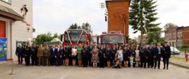 Pamiątkowe zdjęcie strażaków i zaproszonych na uroczystość gości przed czerwonymi strażackimi pojazdami ciężarowymi.