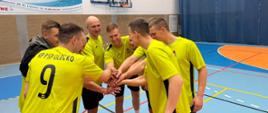 Strażacy z KP PSP w Olecku, ubrani w jasne stroje sportowe, stoją w okręgu i składają ręce przed rozpoczęciem meczu.