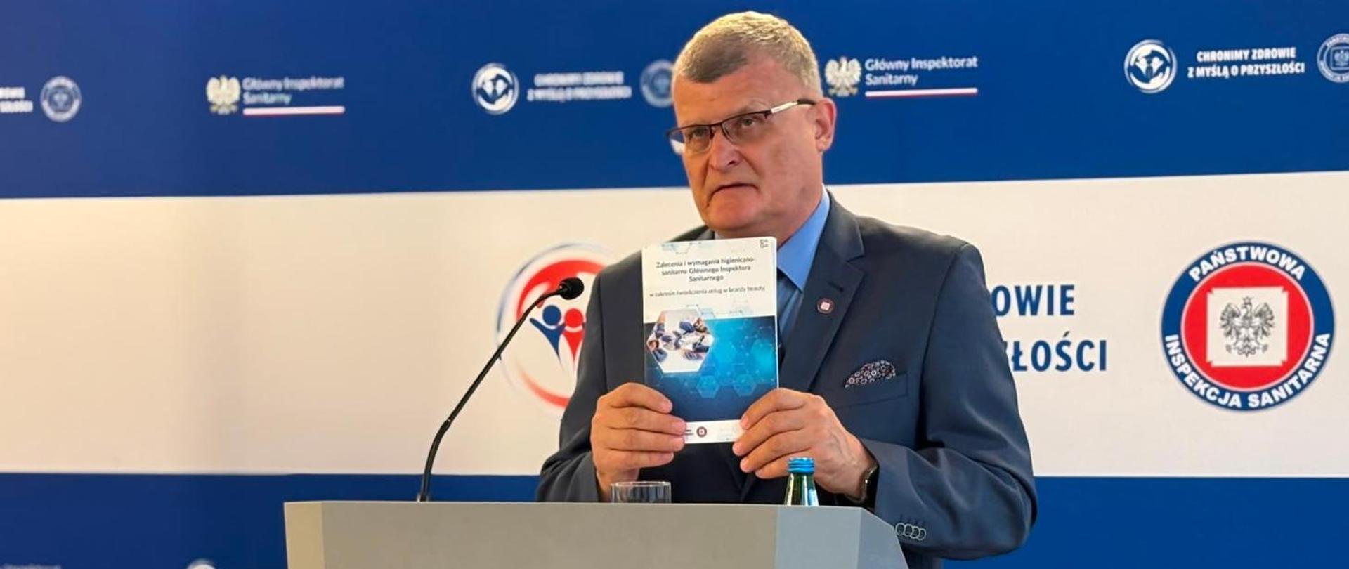 Główny Inspektor Sanitarny przy mównicy podczas konferencji prasowej, z publikacją w rękach