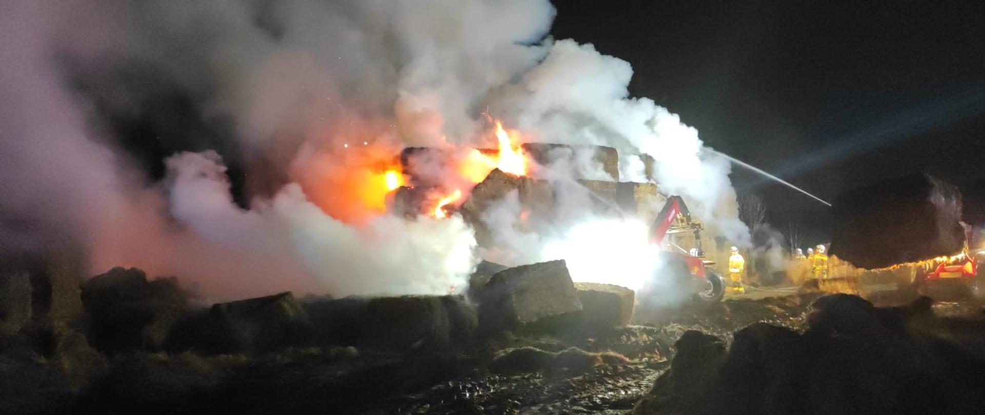 Zdjęcie zrobione nocą. Pali się sterta balotów w dużych kostkach. Widać płomienie i dużo białego dymu.