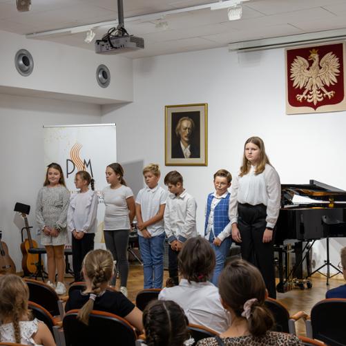 Międzynarodowy Dzień Muzyki 2023. Grupa uczniów, którzy przygotowali koncert oraz pani Agata Kopyto na scenie PSM w Głubczycach. W prawym górnym rogu widoczne godło Polski. W centrum zdjęcia za grupą uczniów widać baner reklamowy szkoły z okazji 50-lecia PSM. Na pierwszym planie u dołu zdjęcia widać publiczność.