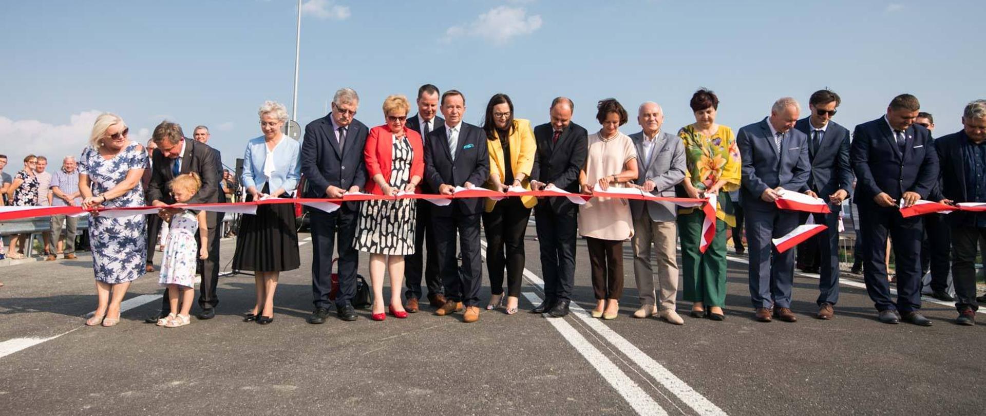 Kilkanaście osób przecina biało-czerwoną wstęgę na nowej drodze.