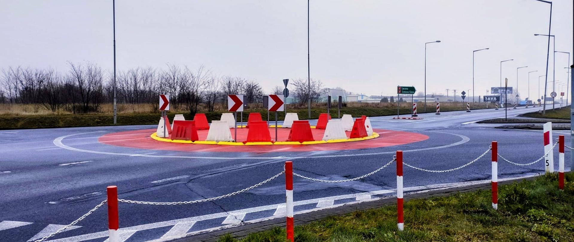 Tymczasowe rondo na drodze, wykonane z czerwono-białych plastikowych barier na czerwonym okręgu z żółtą obwódką, otoczone asfaltową jezdnią i znakami kierunkowymi. W tle widoczne latarnie i pusty teren pod pochmurnym niebem.