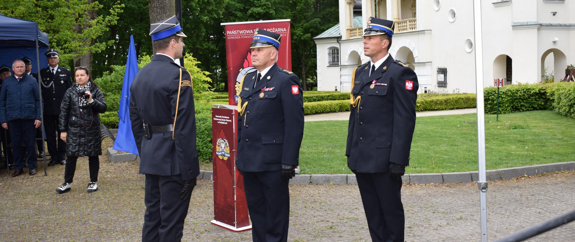 Jeden strażak stoi na baczność naprzeciwko dwóch innych strażaków. Ubrani są w czarne strażacki mundury 