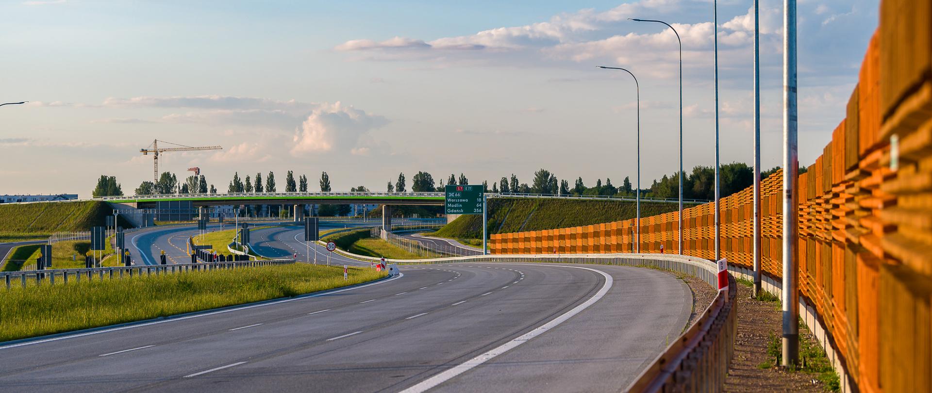 Zdjęcie przedstawia fragment drogi ekspresowej S7, na której widoczna jest szeroka, wielopasmowa jezdnia z wyraźnym oznakowaniem poziomym. Po obu stronach drogi znajdują się barierki ochronne, a po prawej stronie rozciąga się wysoki, brązowo-pomarańczowy ekran akustyczny. W tle widoczny jest wiadukt oraz znaki drogowe, w tym zielony znak informacyjny zawierający odległości do miast. Horyzont wypełniają drzewa, a w oddali można dostrzec dźwig budowlany. Całość jest oświetlona ciepłym światłem zachodzącego słońca.