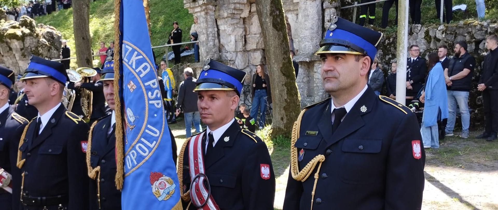 Strażacy Komendy Wojewódzkiej Państwowej Straży Pożarnej w Opolu stoją w galowych mundurach z niebieskim sztandarem PSP. Za nimi widoczne są kamienne ruiny oraz liczni uczestnicy pielgrzymki zgromadzeni na trawiastym zboczu.