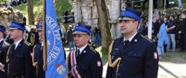 Strażacy Komendy Wojewódzkiej Państwowej Straży Pożarnej w Opolu stoją w galowych mundurach z niebieskim sztandarem PSP. Za nimi widoczne są kamienne ruiny oraz liczni uczestnicy pielgrzymki zgromadzeni na trawiastym zboczu.