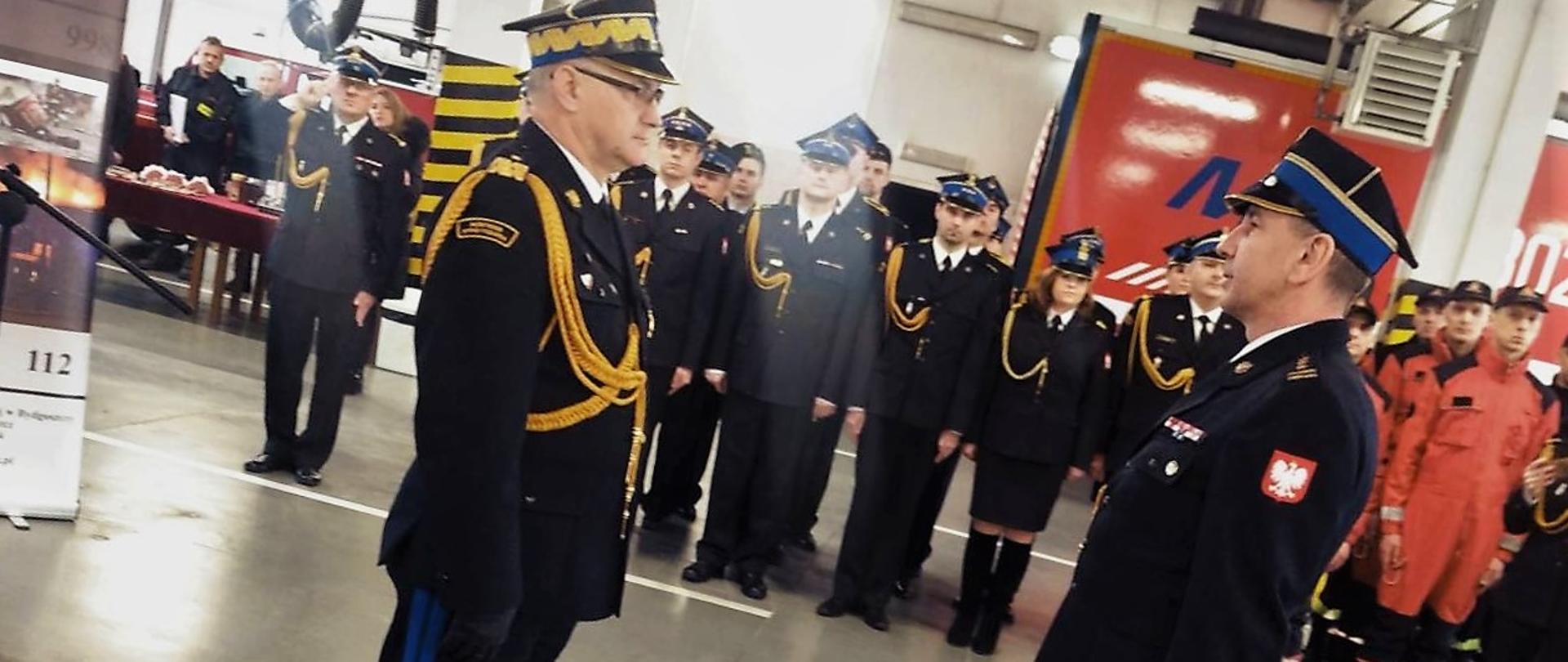 Komendant miejski składa ostatni meldunek przełożonemu. W tle załoga i goście uroczystości. Z boki widać fragment banera. Rzecz dzieje się w garażu.