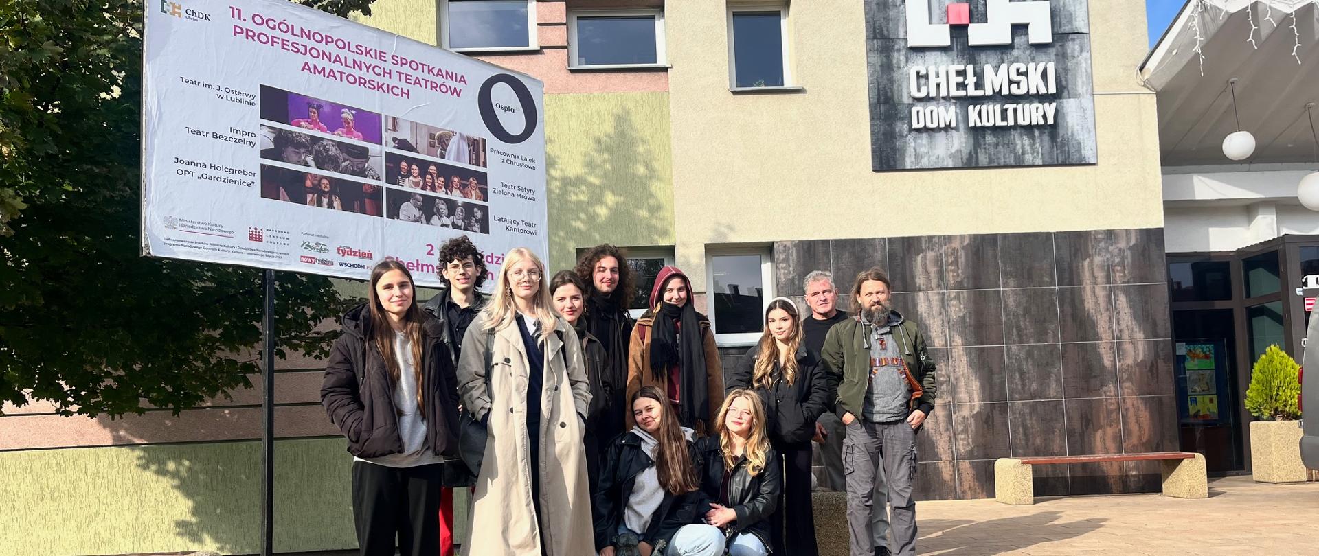 Grupa siedmiu dziewcząt, spośród których dwie przykucają a pięć stoi za nimi, dwóch chłopców stojących za dziewczętami oraz dwóch mężczyzn stojących z prawej strony pozuje do zdjęcia na tle budynku Chełmskiego Domu Kultury. Są ubrani w kurtki i płaszcze. Po prawej stronie widać fragment wejścia do budynku oraz biało - czerwone logo i napis Chełmski Dom Kultury. Po lewej stronie jest bilbord, a nami plakat z napisem 11. OGÓLNOPOLSKIE SPOTKANIA PROFESJONALNYCH TEATRÓW AMATORSKICH Ospta. Poniżej jest sześć fotosów w dwóch rzędach, a obok nich podpisy. Z lewej strony: Teatr im. J. Osterwy w Lublinie; Impro Teatr Bezczelny; Joanna Holcberger OPT "Gardzienice"; Pracownia Lalek z Chrustowa; Teatr Satyry Zielona Mrówa oraz Latający Teatr Kantorowi. Na dole logotypy organizatorów i patronów.