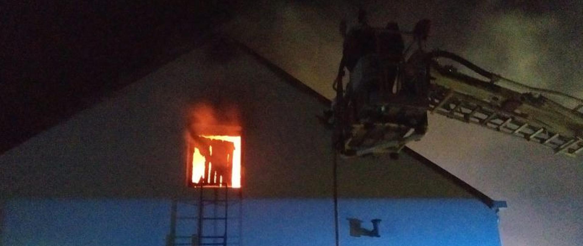 Zdjęcie wykonane w nocy. W centralnym miejscu zdjęcia budynek oświetlony lampami błyskowymi. Budynek ma dwuspadowy dach. U szczytu okienko z którego wydostaje się ogień. Przy budynku stoi podnośnik strażacki