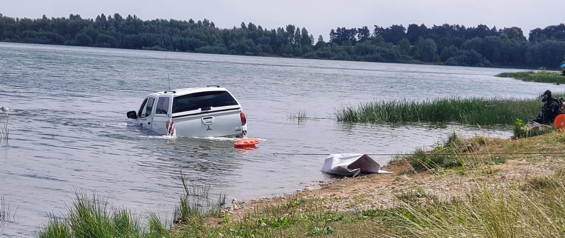 Obraz przedstawia samochód terenowy wyciągany ze zbiornika Słup. Na brzegu siedzi nurek. Pora dzienna