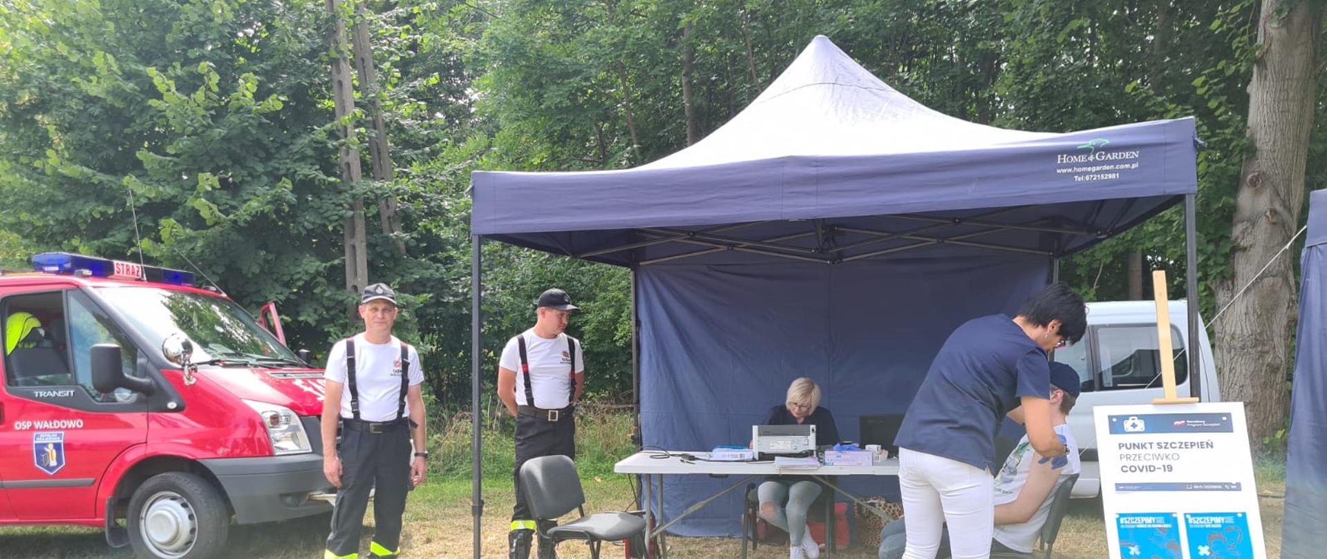 Zdjęcie wykonano porą dzienną, na trawie rozłożony mobilny punkt szczepień, jedna osoba poddaje się szczepieniu, dwóch strażaków Ochotniczej Straży Pożarnej ubranych w mundury koszarowe stoi obok namiotu. Na dalszym planie lekki samochód gaśniczy Ochotniczej Straży Pożarnej w Wałdowie oraz zadrzewienia.