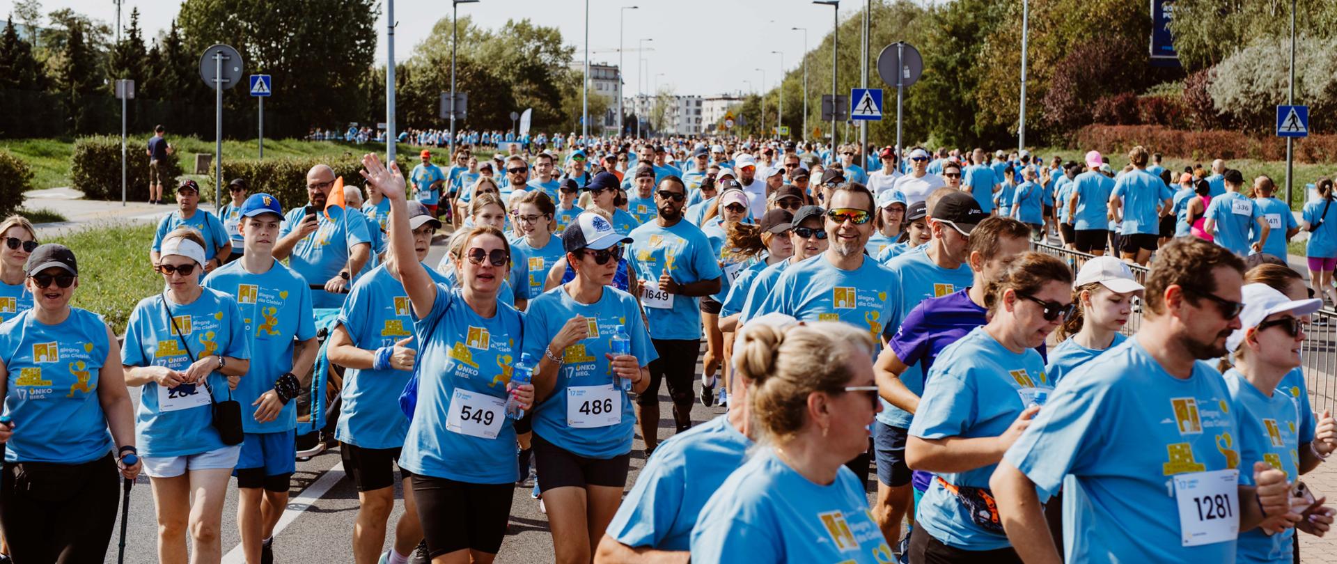 Onkobieg. Tłum ludzi w różnym wieku w niebieskich koszulkach biegnie ulicą. Po bokach roślinność. W tle widać zabudowania.