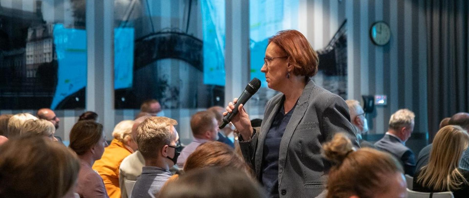 Kobieta z mikrofonem stoi pośród siedzącej widowni, w ręku trzyma wskaźnik laserowy