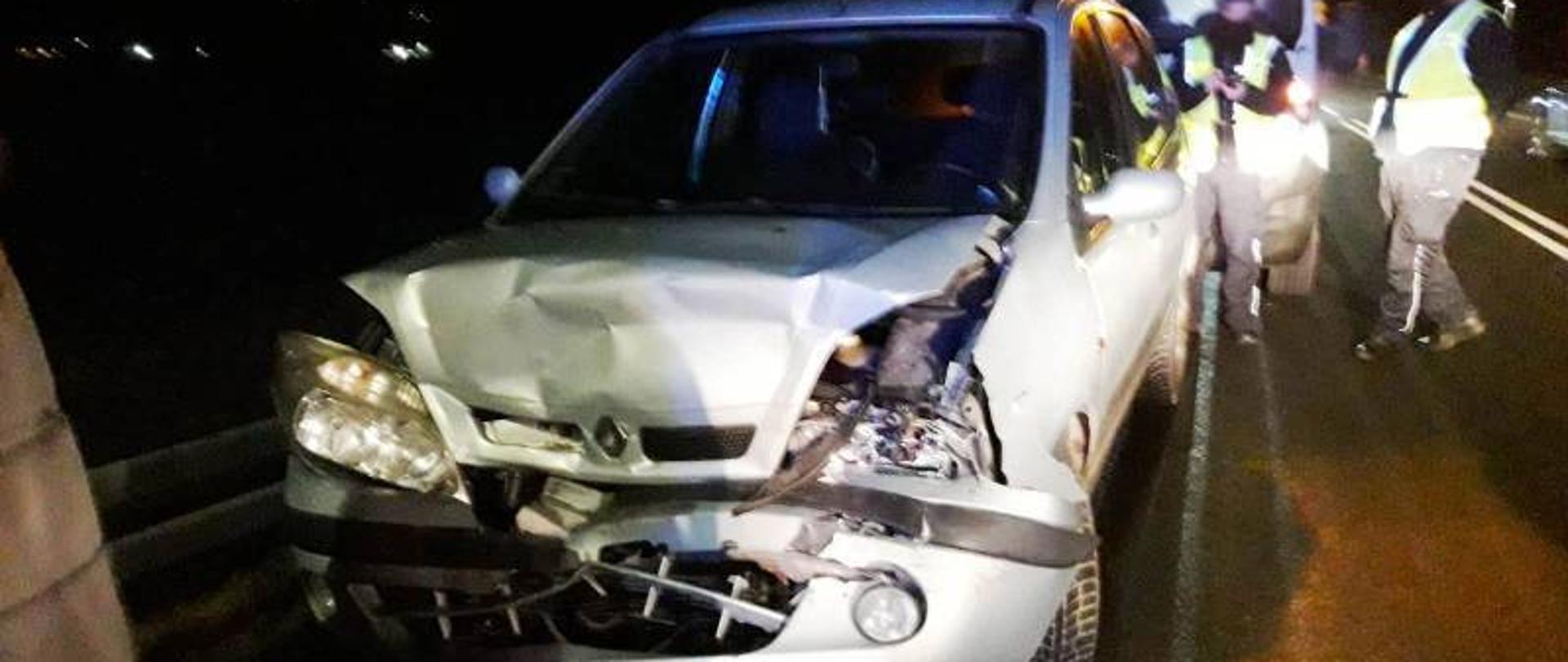 Rozbity przód samochodu uczestniczącego w zdarzeniu w Starym Sączu. Z tyłu stoi inny samochód oraz Policjanci w kamizelkach odblaskowych.