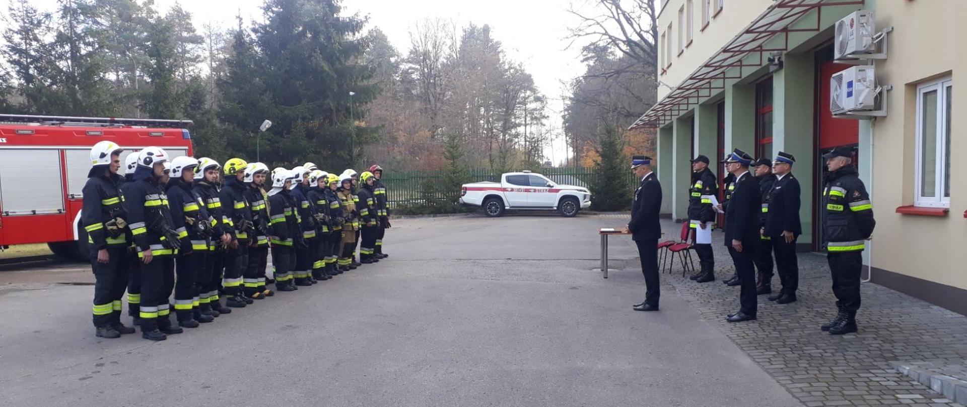 Grupa strażaków w umundurowaniu bojowym stoi przed budynkiem. Naprzeciw grupy stoją inni strażacy, z których jeden zwraca się do grupy. 