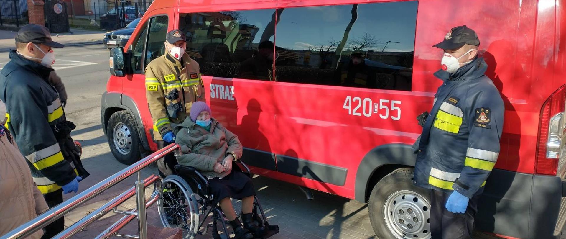 Trzech strażaków w mundurach i maseczkach ochronnych w trakcie transportu osoby starszej na szczepienie. Jeden ze strażaków wiezie kobietę na wózku inwalidzkim. Za nimi widoczny samochód strażacki typu bus koloru czerwonego z przyciemnionymi szybami. Jest pogodnie i słonecznie.