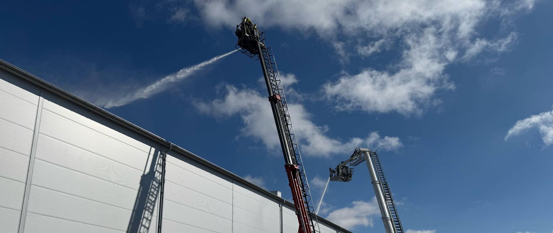 Na zdjęciu dwa podnośniki podają wodę z działek na palący się dach hali produkcyjno-magazynowej.