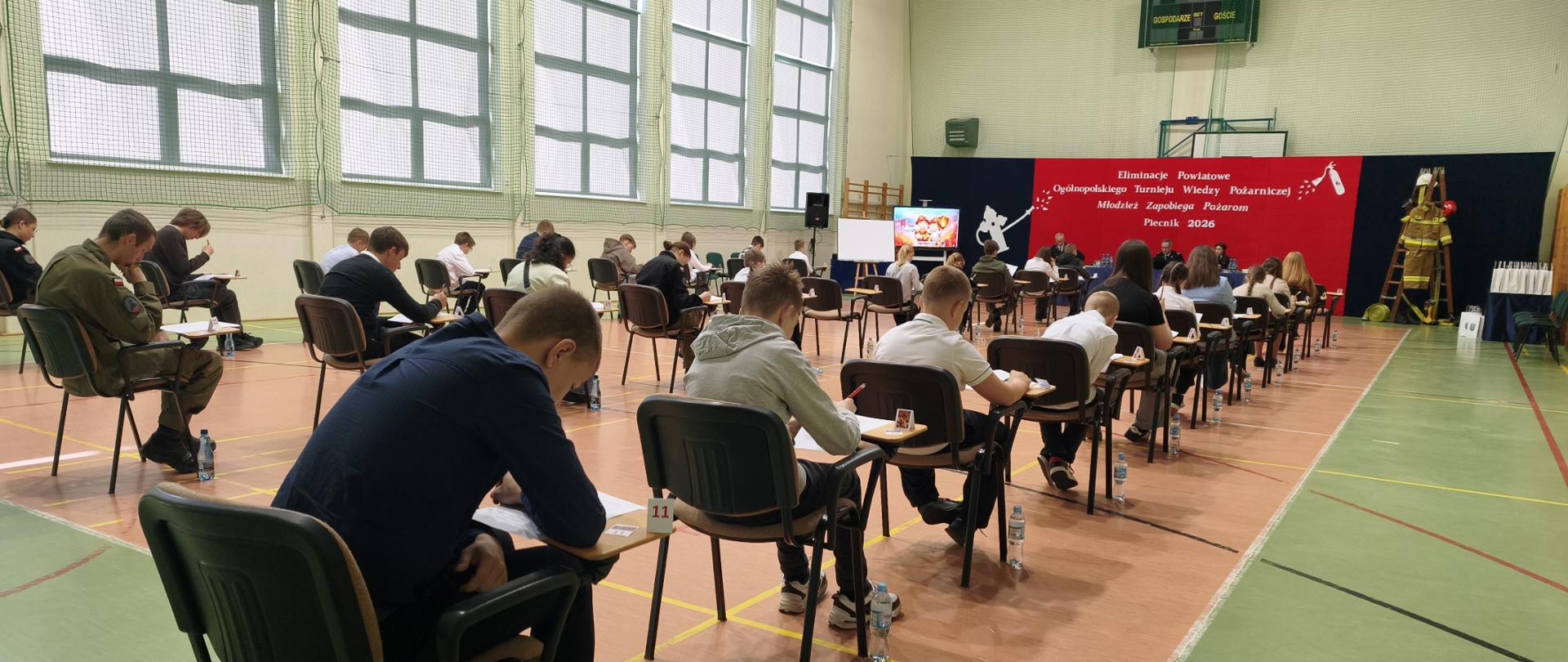Na zdjęciu widoczni uczestnicy konkursu w trakcie pisania testu. Każdy z nich siedzi pochylony na swoją praca w jednoosobowej ławce. Kilkanaście ławek w trzech rzędach sprawia wrażenie wypełnienia sali gimnastycznej