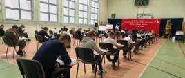 Na zdjęciu widoczni uczestnicy konkursu w trakcie pisania testu. Każdy z nich siedzi pochylony na swoją praca w jednoosobowej ławce. Kilkanaście ławek w trzech rzędach sprawia wrażenie wypełnienia sali gimnastycznej
