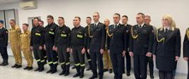 Dwuszereg strażaków na uroczystej zbiórce. Na zdjęciu widocznych około 20 osób.