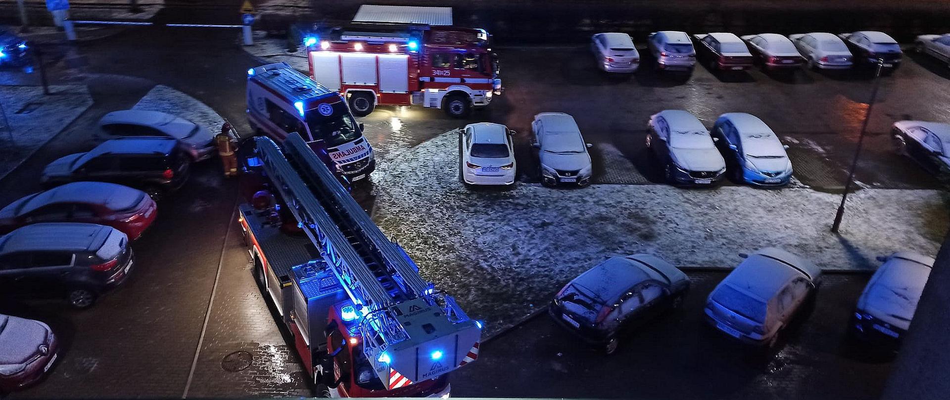 Widok z okna na parking przed budynkiem. Wśród zaparkowanych samochodów osobowych stoją dwa pojazdy pożarnicze i karetka pogotowia z włączonymi światłami alarmowymi.