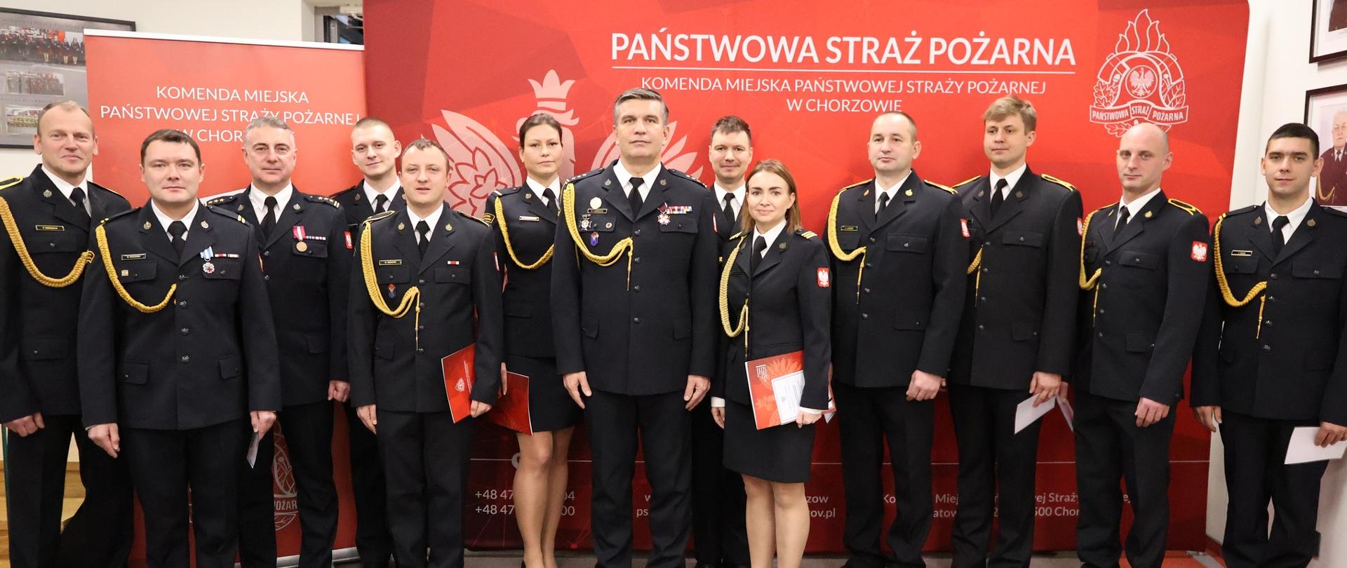 Na tle czerwonego baneru z emblematem Państwowej Straży Pożarnej oraz nazwą Komendy Miejskiej PSP w Chorzowie, grupa funkcjonariuszy w galowych mundurach stoi w równym szeregu. Część z nich trzyma w dłoniach teczki z wręczonymi awansami, co sugeruje moment wręczenia wyróżnień lub podziękowań. Uroczysta atmosfera podkreślona jest powagą postawy strażaków oraz oficjalnym charakterem tła. Zdjęcie dokumentuje zbiórkę z okazji Narodowego Święta Niepodległości – wydarzenie łączące tradycję, służbę i patriotyzm.
