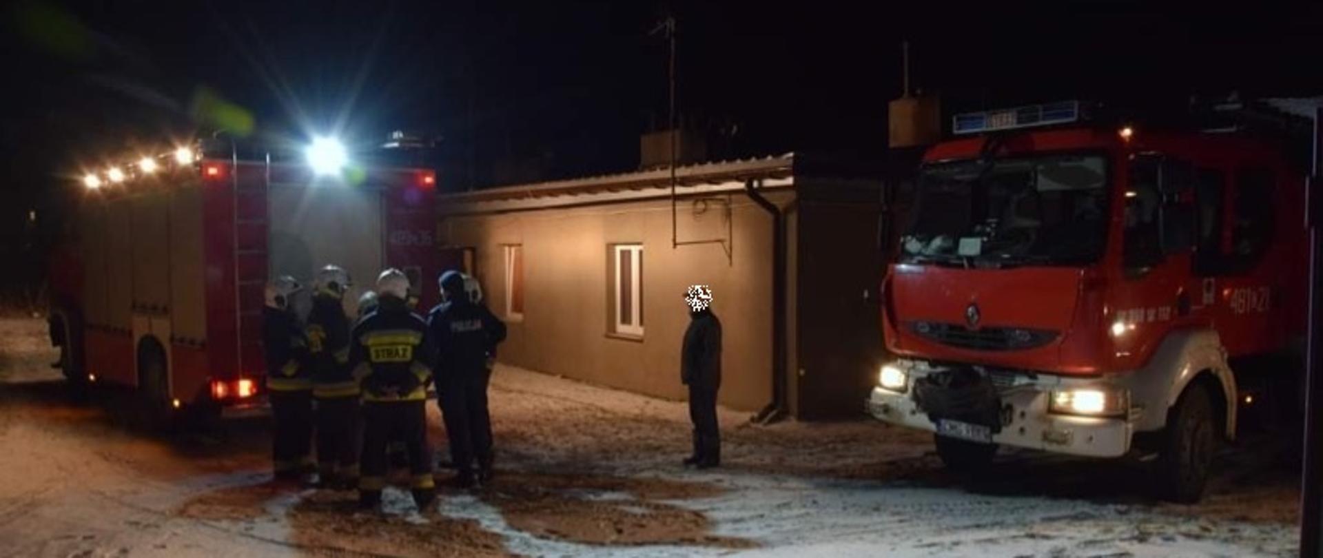 Po środku budynek mieszkalny. Po lewej stronie stoją strażacy za samochodem pożarniczym, z prawej strony drugi samochód pożarniczy